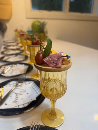 Mini charcuterie appetizer with salami, cherry grape, basil and cheese skewer on a wooden lid perched atop an amber vintage goblet, arranged in a row on a stylish indoor dinner table setting.