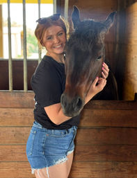 Smiling woman with sunglasses on her head hugging a dark bay horse inside a sunlit wooden stable stall