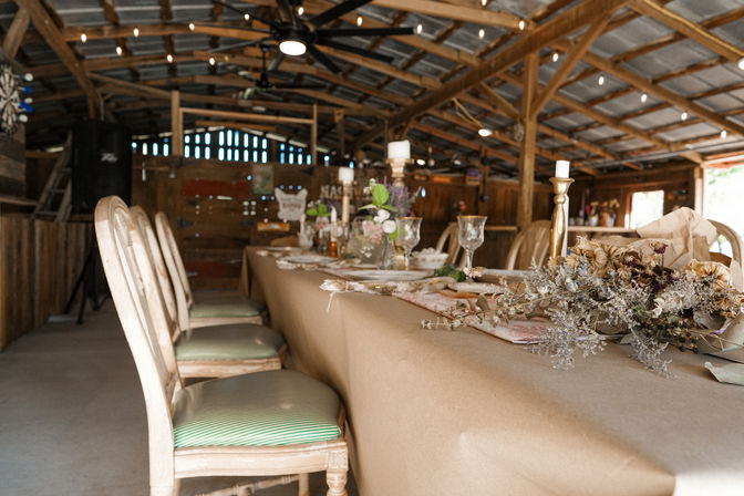Cozy rustic barn dining table set for a celebration with mismatched wooden chairs, kraft-paper table covering, glassware, brass candlesticks, and dried floral centerpieces beneath exposed wooden beams and string lights.