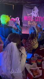 Bachelorette group watching a lively dueling drag show in a dim nightclub, bride wearing a white veil, neon-green wig performer on stage, rainbow party cups and snacks on the table.
