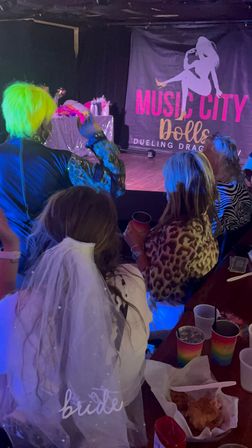 Bachelorette group watching a lively dueling drag show in a dim nightclub, bride wearing a white veil, neon-green wig performer on stage, rainbow party cups and snacks on the table.