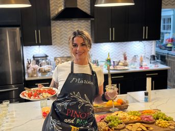 Host in a modern home kitchen holding a plate of caprese salad and a wine glass at a marble island with a large charcuterie board of cheeses, grapes, crackers and cured meats under pendant lights
