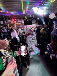 Crowded indoor bar with colorful streamers and disco lights, performer in a silver fringe cowgirl outfit and white hat kneeling in the aisle while patrons at tables cheer and film on phones