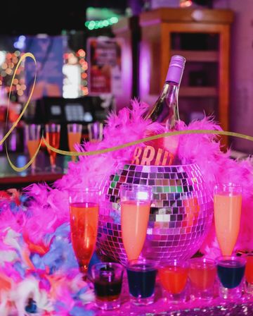 Vibrant nightclub table: disco-ball ice bucket holding a rosé bottle draped in a hot-pink feather boa, surrounded by colorful cocktails and shot glasses under neon lights