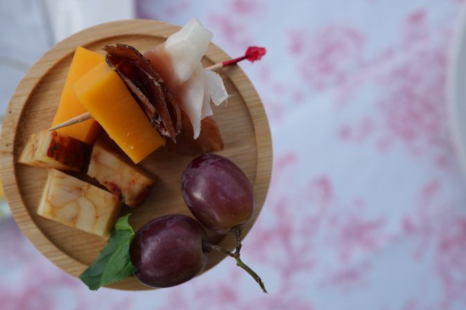 Close-up of a small wooden charcuterie plate with cheddar and marbled cheese cubes, cured meat skewered on a toothpick, and two purple grapes on a pink floral tablecloth