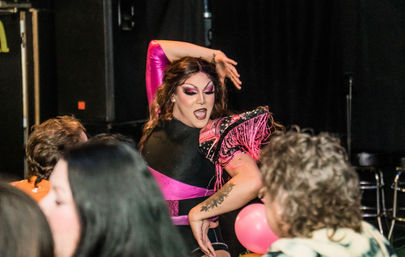 Drag performer in pink-and-black costume with bold makeup striking a dramatic pose during a lively drag show at a nightclub, with blurred audience members and pink balloons in the foreground.