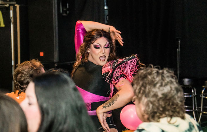 Drag performer in pink-and-black costume with bold makeup striking a dramatic pose during a lively drag show at a nightclub, with blurred audience members and pink balloons in the foreground.