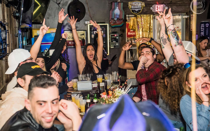 Lively downtown bar scene at night with a mixed group cheering around a crowded table of drinks, hands raised, tattoos visible, neon signs and an EXIT sign in the background — energetic city nightlife.