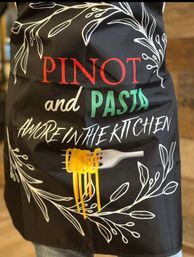 Black kitchen apron with white botanical illustrations, bold red PINOT and green PASTA lettering, a fork twirling yellow spaghetti and the handwritten phrase amore in the kitchen