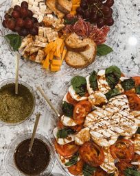 Overhead appetizer spread on a marble countertop: a caprese salad of sliced mozzarella, tomatoes, basil and balsamic drizzle, and a charcuterie platter with cubed and sliced cheeses, salami, grapes, crostini plus bowls of pesto and dressing.