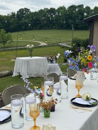 Rustic outdoor event tablescape on a countryside lawn with white linen, amber goblets, floating candle cylinders, colorful wildflower centerpieces and string lights overlooking rolling green fields.