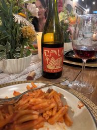 Cozy candlelit Italian dinner scene: a plate of tomato pasta with fork in foreground, a bottle reading Terre Siciliane and a glass of red wine on a rustic wooden table next to a floral centerpiece and cork.