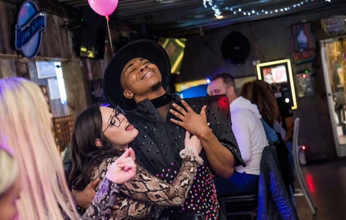Lively bar scene with two people dancing and singing — one in a wide-brim black hat and sequined outfit leaning back with hand on chest, the other in glasses and a snake-print top hugging them, colorful lights and a pink balloon overhead.