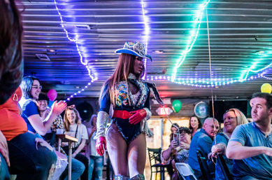 Drag performer in a sequined cowboy hat and glittery bodysuit walks through a lively indoor bar, neon string lights overhead as a cheering crowd claps and records on phones.