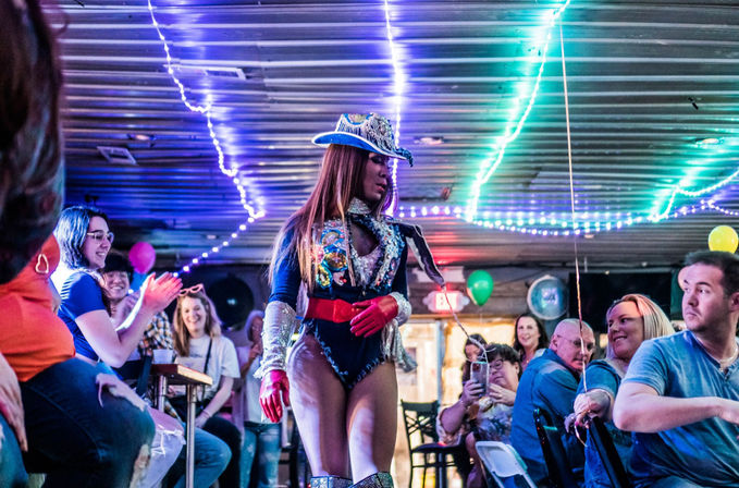 Drag performer in a sequined cowboy hat and glittery bodysuit walks through a lively indoor bar, neon string lights overhead as a cheering crowd claps and records on phones.