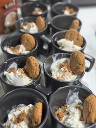 Rows of black ceramic mugs filled with vanilla ice cream, sprinkled with cookie crumbs and each garnished with a whole spiced cookie — bite-sized dessert cups ready to serve.