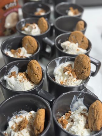 Rows of black ceramic mugs filled with vanilla ice cream, sprinkled with cookie crumbs and each garnished with a whole spiced cookie — bite-sized dessert cups ready to serve.