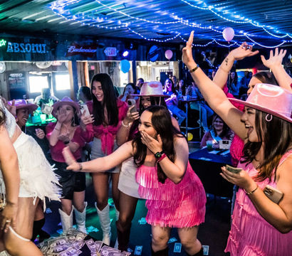 Bachelorette party at a lively country bar — women in pink fringe dresses and cowboy hats cheer and toss dollar bills toward a dancer on stage under string lights