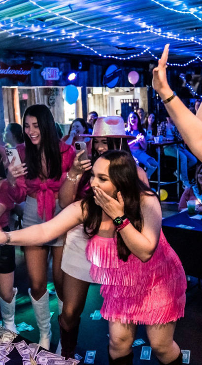 Bachelorette party at a lively country bar — women in pink fringe dresses and cowboy hats cheer and toss dollar bills toward a dancer on stage under string lights
