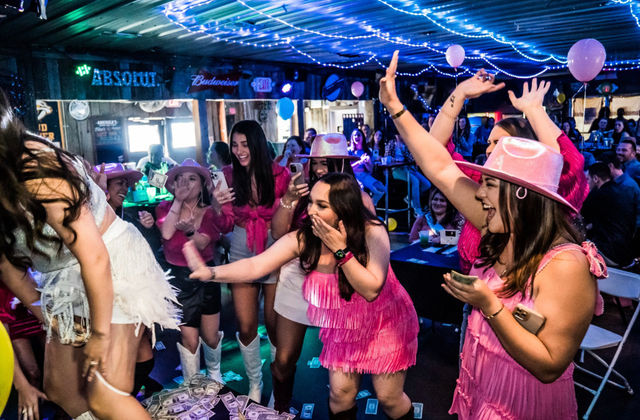 Bachelorette party at a lively country bar — women in pink fringe dresses and cowboy hats cheer and toss dollar bills toward a dancer on stage under string lights