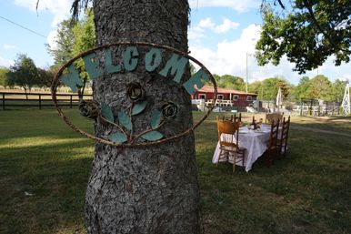 Rustic metal 'WELCOME' sign with rose motifs mounted on a tree in a country yard, long outdoor dining table with mismatched wooden chairs set on the lawn near a red barn and fence under a blue sky