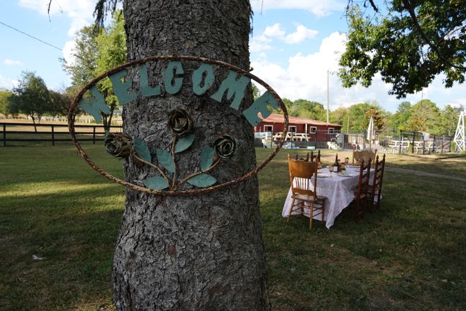 Rustic metal 'WELCOME' sign with rose motifs mounted on a tree in a country yard, long outdoor dining table with mismatched wooden chairs set on the lawn near a red barn and fence under a blue sky