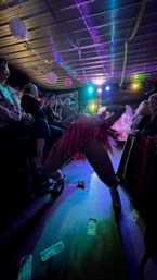 Drag performer in pink fringe costume leaning back on a small club stage as cheering audience members film and clap, neon spotlights, string lights and paper lanterns overhead, dollar bills scattered on the floor.
