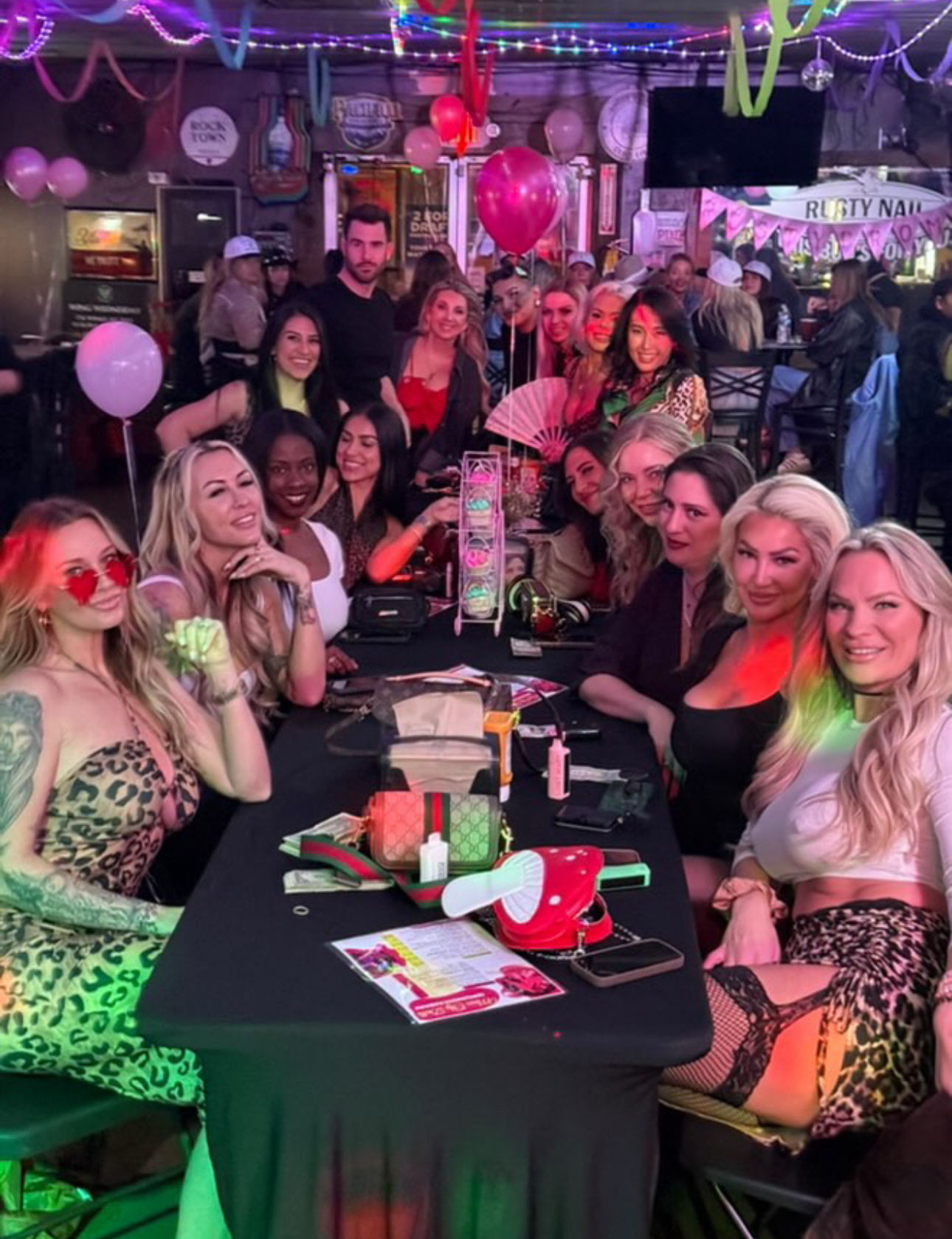 Large group of women enjoying a girls' night out at a lively indoor bar — long table with purses, drinks, pink balloons, neon string lights and party decorations.