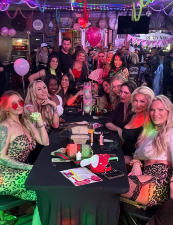 Large group of women enjoying a girls' night out at a lively indoor bar — long table with purses, drinks, pink balloons, neon string lights and party decorations.