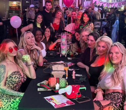 Large group of women enjoying a girls' night out at a lively indoor bar — long table with purses, drinks, pink balloons, neon string lights and party decorations.