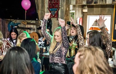 Group of women enjoying a girls' night out, dancing and cheering with hands raised in a rustic country-style bar with wood-paneled walls, guitars on the wall and a pink balloon.