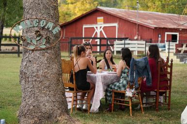 Rustic farm scene: a group of women enjoying outdoor dining at a cloth-covered table under a tree with a metal “Welcome” sign, with a red barn and fenced pasture in the background.