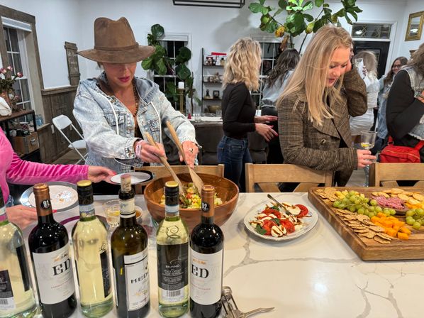 Guests at a lively indoor gathering around a kitchen island — a woman in a wide‑brimmed hat mixes a large wooden bowl of salad while others hold wine glasses and help themselves to a charcuterie board with cheeses, crackers, grapes and sliced tomatoes.