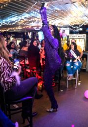 Drag performer in a sequined purple costume strikes a dramatic pose in a string-lit bar while a lively crowd watches and records the show on phones.