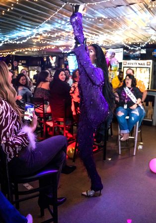 Drag performer in a sequined purple costume strikes a dramatic pose in a string-lit bar while a lively crowd watches and records the show on phones.