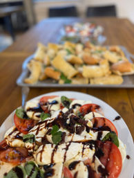 Caprese salad of sliced tomatoes, fresh mozzarella and basil leaves drizzled with balsamic glaze on a white plate, with a blurred tray of bread and crostini on a wooden table