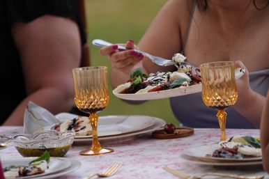 Al fresco summer dinner close-up — person serving Caprese salad with fresh mozzarella, tomatoes, basil and balsamic glaze, amber goblets and plates on a patterned tablecloth.