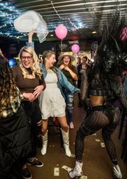 Lively indoor bar scene with string lights and pink balloons — friends cheering as one in white cowboy boots and a white hat tosses, while a dancer in a sparkly sheer outfit performs and dollar bills scatter on the floor.
