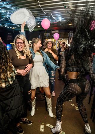 Lively indoor bar scene with string lights and pink balloons — friends cheering as one in white cowboy boots and a white hat tosses, while a dancer in a sparkly sheer outfit performs and dollar bills scatter on the floor.