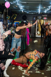 Lively indoor bar drag show with a performer in red thigh-high boots sprawled on the floor, audience cheering and tossing dollar bills, string lights overhead and a pink balloon.