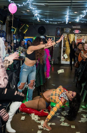 Lively indoor bar drag show with a performer in red thigh-high boots sprawled on the floor, audience cheering and tossing dollar bills, string lights overhead and a pink balloon.