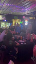 Bachelorette party at a dim bar — group of women around a drink-filled table under string lights and neon signs, pink balloon and a costumed performer with a bright wig entertaining the crowd.
