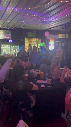 Bachelorette party at a dim bar — group of women around a drink-filled table under string lights and neon signs, pink balloon and a costumed performer with a bright wig entertaining the crowd.