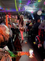 Lively indoor bar scene with a sequined performer in a fringe hat and thigh-high boots walking down the center aisle past seated patrons under colorful streamers and string lights, while an audience member films on a phone and dollar bills lie on the floor.