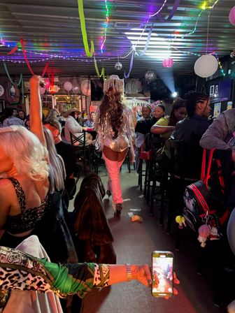 Lively indoor bar scene with a sequined performer in a fringe hat and thigh-high boots walking down the center aisle past seated patrons under colorful streamers and string lights, while an audience member films on a phone and dollar bills lie on the floor.