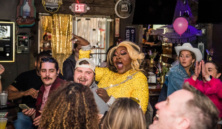 Energetic drag performer in a yellow sequined dress entertains a lively bar crowd during a drag show, guests laughing, cheering and clapping.