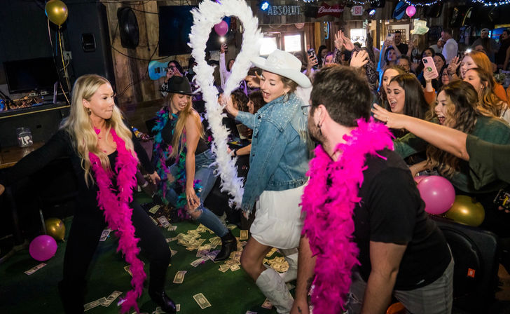 Partygoers dancing at a lively country bar night out, wearing pink feather boas and cowboy hats, cash scattered on the floor, balloons and phones capturing the fun.
