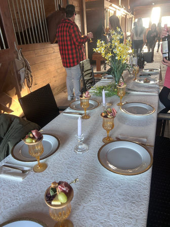 Rustic barn dining table set for a gathering — white lace tablecloth, gold-rimmed plates, amber goblets with fruit and charcuterie skewers, crystal candles and a yellow daffodil centerpiece, guests mingling by wooden stalls.