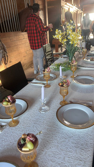 Rustic barn dining table set for a gathering — white lace tablecloth, gold-rimmed plates, amber goblets with fruit and charcuterie skewers, crystal candles and a yellow daffodil centerpiece, guests mingling by wooden stalls.