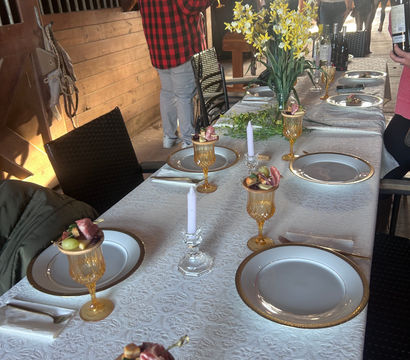 Rustic barn dining table set for a gathering — white lace tablecloth, gold-rimmed plates, amber goblets with fruit and charcuterie skewers, crystal candles and a yellow daffodil centerpiece, guests mingling by wooden stalls.